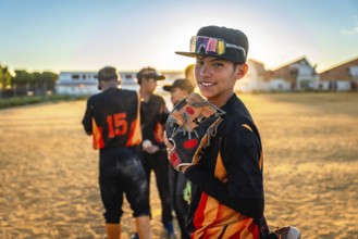 Young baseball player standing on a dirt field, holding a glove, smiling at the camera during a