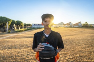 Young baseball player stands on a dusty field at golden hour, wearing a black uniform and cap,