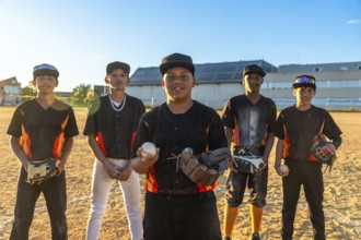 Group of diverse teenage baseball players standing on a dusty field, one holding a ball and glove,