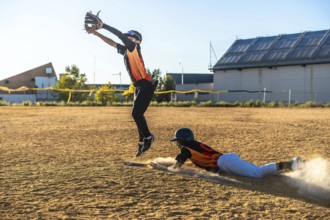Baseball player is sliding into base creating a dust cloud while a defensive player is jumping high