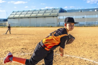 Young boy in baseball uniform and cap delivering a pitch from the mound on a sunlit dirt field,