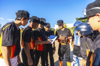 Baseball coach in black and orange uniform reviews strategy on a clipboard with attentive youth
