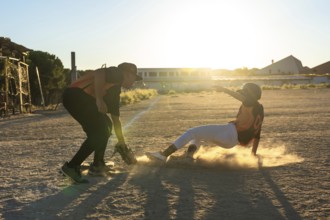 Baseball player sliding into a base, creating a cloud of dust, with another player preparing to