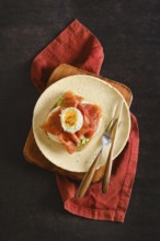 Top view of toasted bun with avocado, cheese topped with smoked salmon and poached egg. Utensils