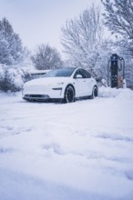 White electric car standing in a snowy landscape next to a charging station, Tesla Model Y electric