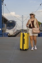 Middle aged woman standing on a railway platform with a bright yellow suitcase, waiting for someone