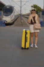 Adult female traveler standing on a railway platform with a yellow suitcase, looking around while