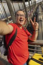 Middle aged man wearing reading glasses smiles with teeth while taking a selfie and making a peace