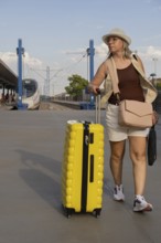 Adult woman traveling alone at a city train station, pulling a yellow suitcase while walking along