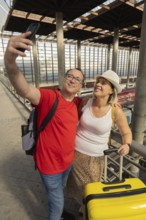 Smiling adult travel couple taking a selfie and looking at a smartphone while standing inside a