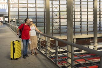 Smiling adult travel couple standing with luggage on an upper level of a modern train station,