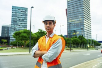 Confident man wearing a hard hat and safety vest standing with crossed arms in a modern urban area,