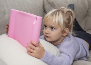 Portrait of a blonde girl, 4 years old, lying down reading a tablet in Ystad, Skåne County, Sweden,
