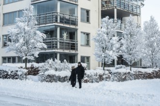 A couple walks on a snow-covered street in front of apartment buildings and snow-laden trees in