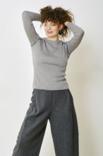 A woman stands with her hands behind her head, showcasing a cozy grey long-sleeve top paired with
