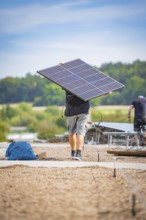 A man carries a solar panel on a construction site with a wooded background, new park and ride