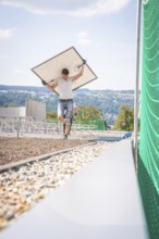 Man carrying a solar panel across a building site with a view and green fence, new park and ride