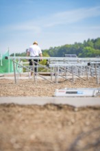 A worker assembles solar panel structures on a gravel roof under a blue sky, new park and ride
