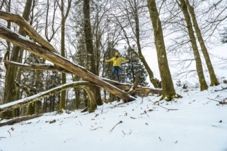 Man wearing a yellow jacket and hat balancing arms outstretched on a large fallen tree log covered