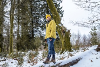 Man wearing a yellow jacket and beanie standing on a fallen tree trunk in a snowy forest, enjoying