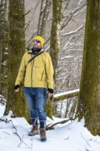 Man wearing a yellow winter jacket, jeans, and hiking boots exploring a snowy forest landscape,