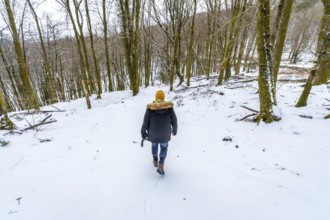 Man in a winter coat and yellow hat walking away into a snow covered forest, exploring and enjoying