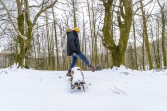 Man wearing winter clothing and a yellow beanie balancing on a snow covered log, enjoying the cold