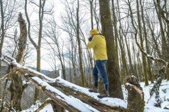 Man wearing a yellow jacket and beanie standing on a snow covered fallen tree, focusing a camera to