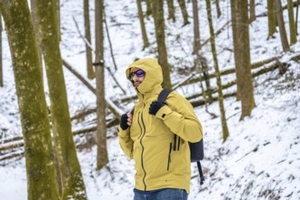 Man in yellow hooded jacket and backpack standing in snowy forest, exploring winter trails and