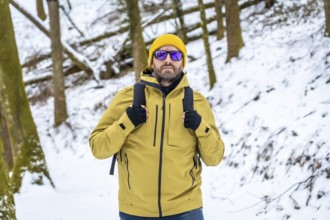 Man in yellow jacket and beanie with sunglasses and backpack hiking through a snow covered forest