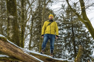Man in yellow jacket and beanie standing on a fallen log in a snowy forest, confidently taking in a