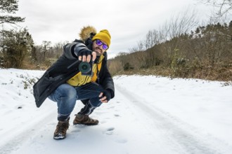 Man in winter gear with a yellow hat and sunglasses crouching on a snowy trail, aiming a camera to