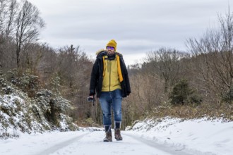 Man in warm winter attire and sunglasses walking on a snowy road through a woodland landscape,
