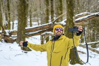 Man wearing a yellow jacket and beanie capturing moments with his camera in a snow covered forest,