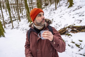 Man wearing an orange beanie and winter jacket, holding a hot mug while contemplating the serene