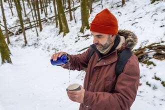 Man wearing an orange beanie and winter jacket poring a hot drink from a blue thermos into a mug,