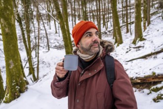 Bearded man in an orange beanie and jacket sips from a mug while standing in a snow covered forest,