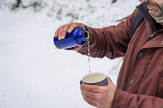 Man wearing a winter jacket pouring a hot drink from a blue thermos bottle into a cup, staying