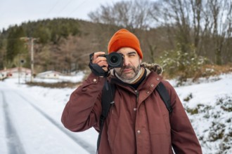 Man in winter clothing and an orange beanie holding a mirrorless camera, capturing nature and