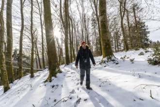 Man walking a snowy forest trail, sunlight filtering through bare trees as he hikes in warm gear,