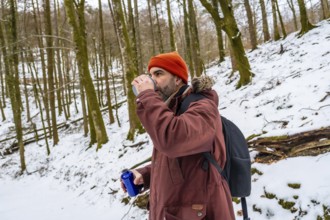 Man in orange beanie and winter jacket drinks from a reusable bottle on a snowy forest trail during