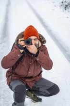 Photographer wearing an orange beanie and parka kneeling in the snow, capturing images with a
