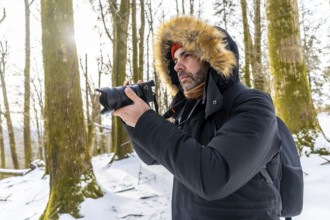 Man in winter parka with fur hood photographing a snowy forest trail, capturing nature with a large