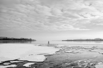 A person walks on a frozen lake under a cloudy sky in a wintry landscape, black and white