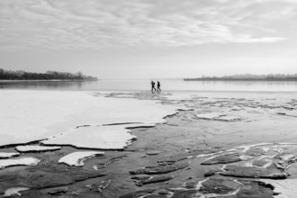 Two people walking on a frozen lake under a cloudy sky in a wintry landscape, black and white