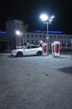 Parking lot at night with white Tesla and charging stations under city lights, Tesla Supercharger,