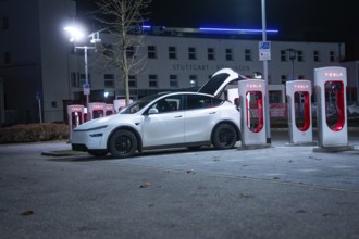Parking lot with white Tesla at night, open rear, charging stations in the background, Tesla