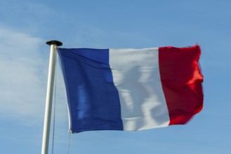 A French flag flutters in the wind on a white flagpole. The background is a clear, azure sky