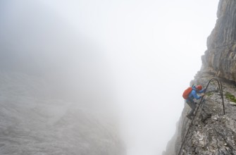 Mountaineer climbs a ladder on the Via Ferrata Oliva Detassis via ferrata in fog, cloudy steep rock