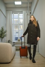 A woman walks through a hallway carrying a red suitcase. She is dressed in a long coat and tall